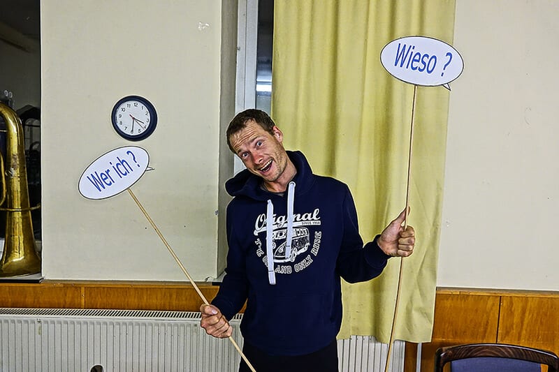 Heiko Gärtner steht in einem Seminarraum und hält zwei Schilder hoch mit der Aufschrift "Wer ich?" - "Wieso?"
